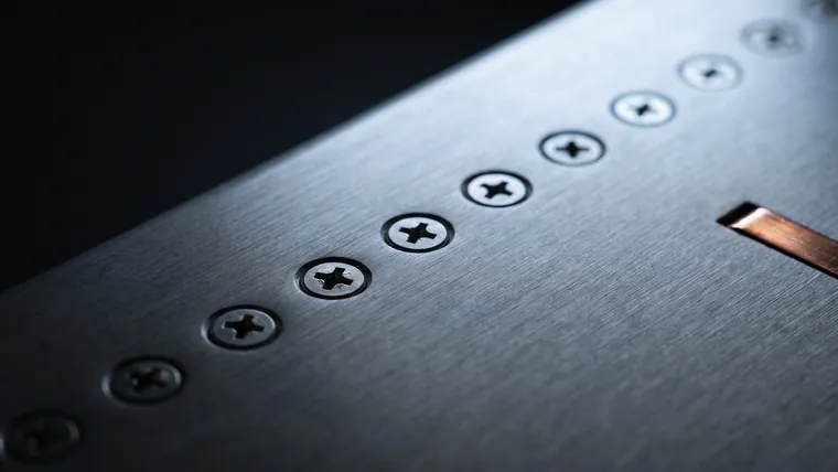 A row of identical steel machine screws driven flush into a brushed metal panel, each seated to the same depth.